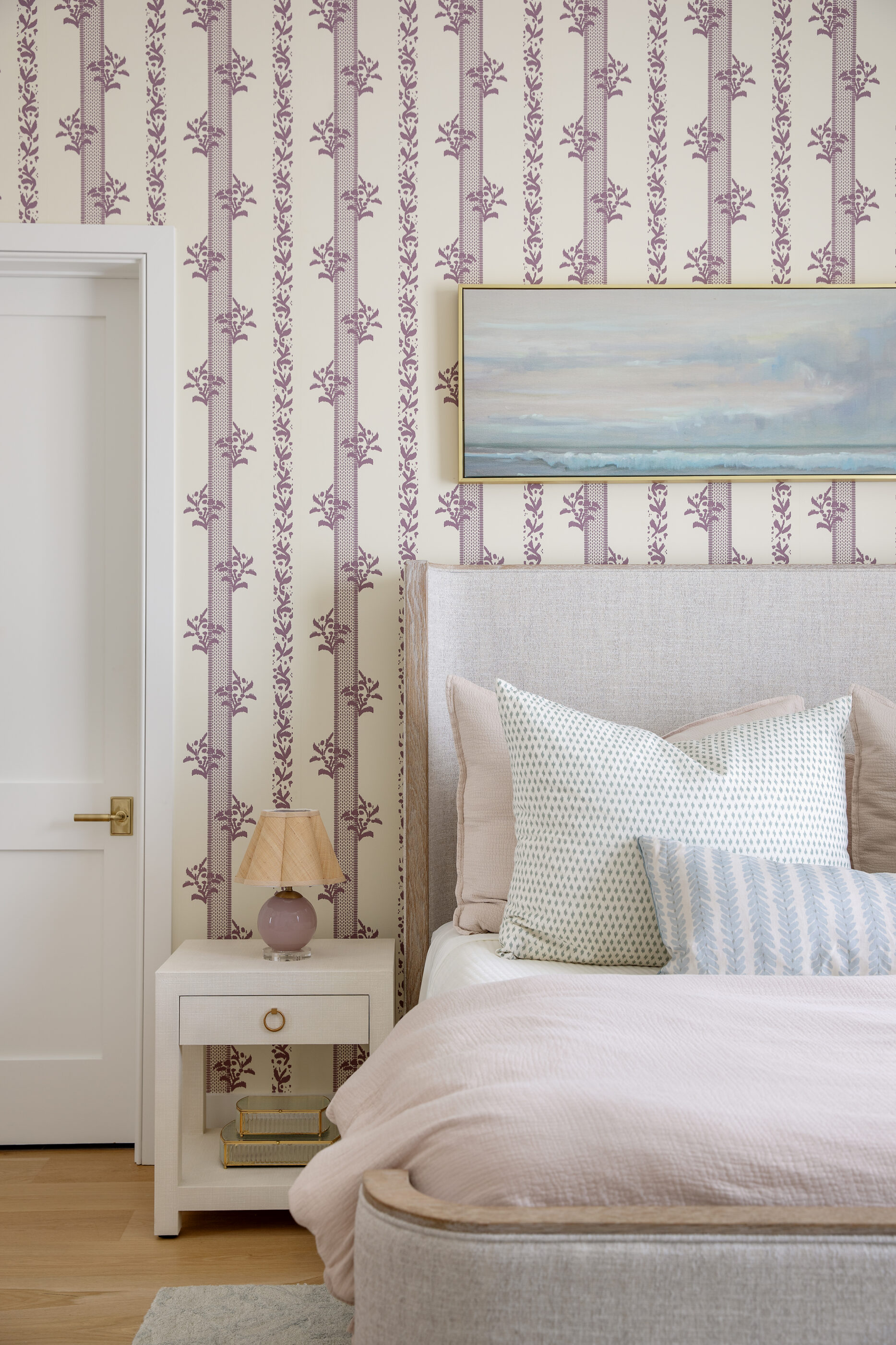 Soft pink and purple bedroom featuring floral striped wallpaper, a textured upholstered bed, layered decorative pillows, coastal artwork, and a white nightstand.